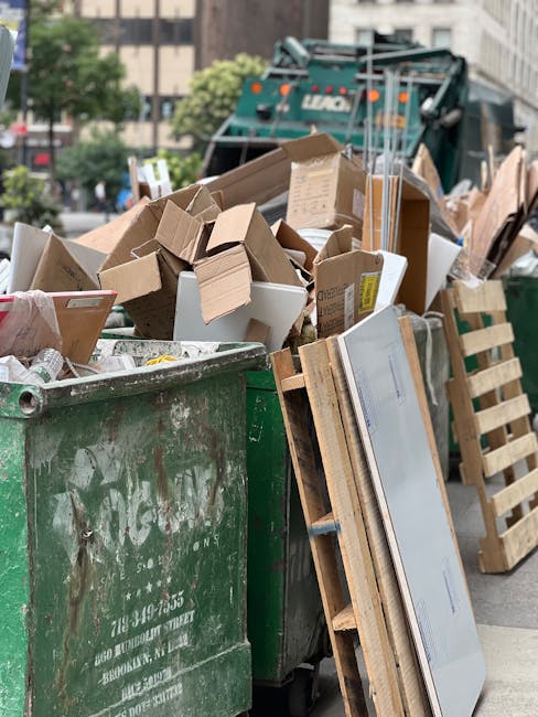 Two waste collection workers are engaged in rubbish removal in an urban environment, standing beside a large waste bin mounted on a compact garbage truck. The worker on the left, wearing a dark green 