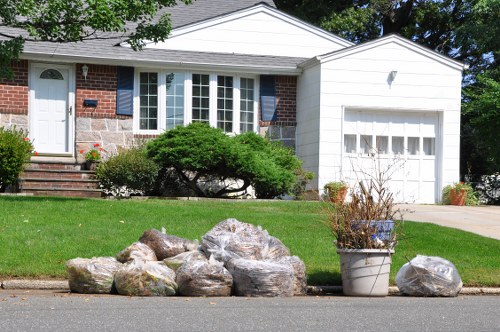 In front of a single-story suburban house with a white garage door and a brick exterior, there is a collection of large, irregularly shaped rocks placed directly on the curbside pavement. To the right of the rocks, a potted plant with bare branches is situated on the pavement. The house features a bay window with white frames and is surrounded by a neatly maintained lawn with a small, dense green shrub in front. The house's steps lead up to a white front door with a small window and a brick pathway runs alongside the entrance. The scene appears to be outdoors on a bright, sunny day, with a backdrop of trees and a partly cloudy sky, illustrating a typical residential area where a local rubbish removal service like Business Waste Removal Blackheath may assist with clearing excess garden and construction debris for properties in the area.