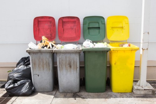 The image displays four large wheelie bins positioned side by side on a paved outdoor surface against a white wall, with the lids open and various types of rubbish visible inside. From left to right, the first bin has a grey body with a red lid, filled with mixed waste including paper, plastic containers, and packaging materials; next, the second bin is also grey with its red lid open, containing similar general waste. The third bin features a green body with its green lid open, holding assorted rubbish, while the fourth bin is yellow with its yellow lid slightly ajar, also filled with waste. To the far left, there are several black plastic rubbish bags stacked against the grey bin. The setting suggests a typical waste collection point on a property exterior, possibly on a driveway or pavement area, with natural daylight illuminating the scene. Business Waste Removal Blackheath performs rubbish management services in this area, supporting local waste disposal needs effectively, as indicated by the surroundings and type of waste depicted. The environment appears clean and organized, with no visible signage or additional equipment present.