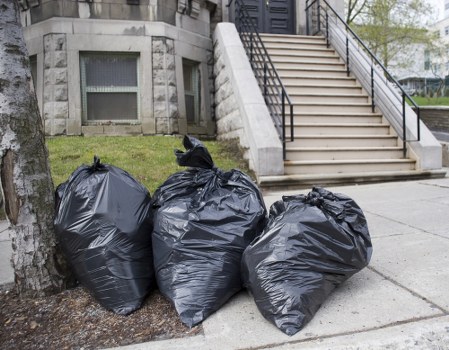 Three large black rubbish bags made from glossy plastic are placed on the sidewalk near a tree trunk in front of a building with stone steps and black metal handrails leading up to an entrance. The bags appear to contain household waste or general rubbish, with some tied at the top and bulging slightly from their contents. The setting is an urban area, possibly in Blackheath or a nearby district, with a paved pavement and a grassy patch beside the tree. The building in the background has a stone façade with small windows at ground level, and the overall scene is illuminated by natural daylight, suggesting daytime hours. Business Waste Removal Blackheath offers professional rubbish removal services in the area, and these bags likely represent waste picked up from residential or commercial properties in the locality.